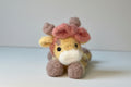 A handmade plush toy depicting a yellow giraffe with a pink bow on its head, lying down against a neutral background.